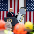 Political speaker at podium with American flags, addressing audience of workers in safety helmets