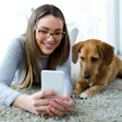 Smiling woman in glasses using smartphone next to golden retriever on carpet