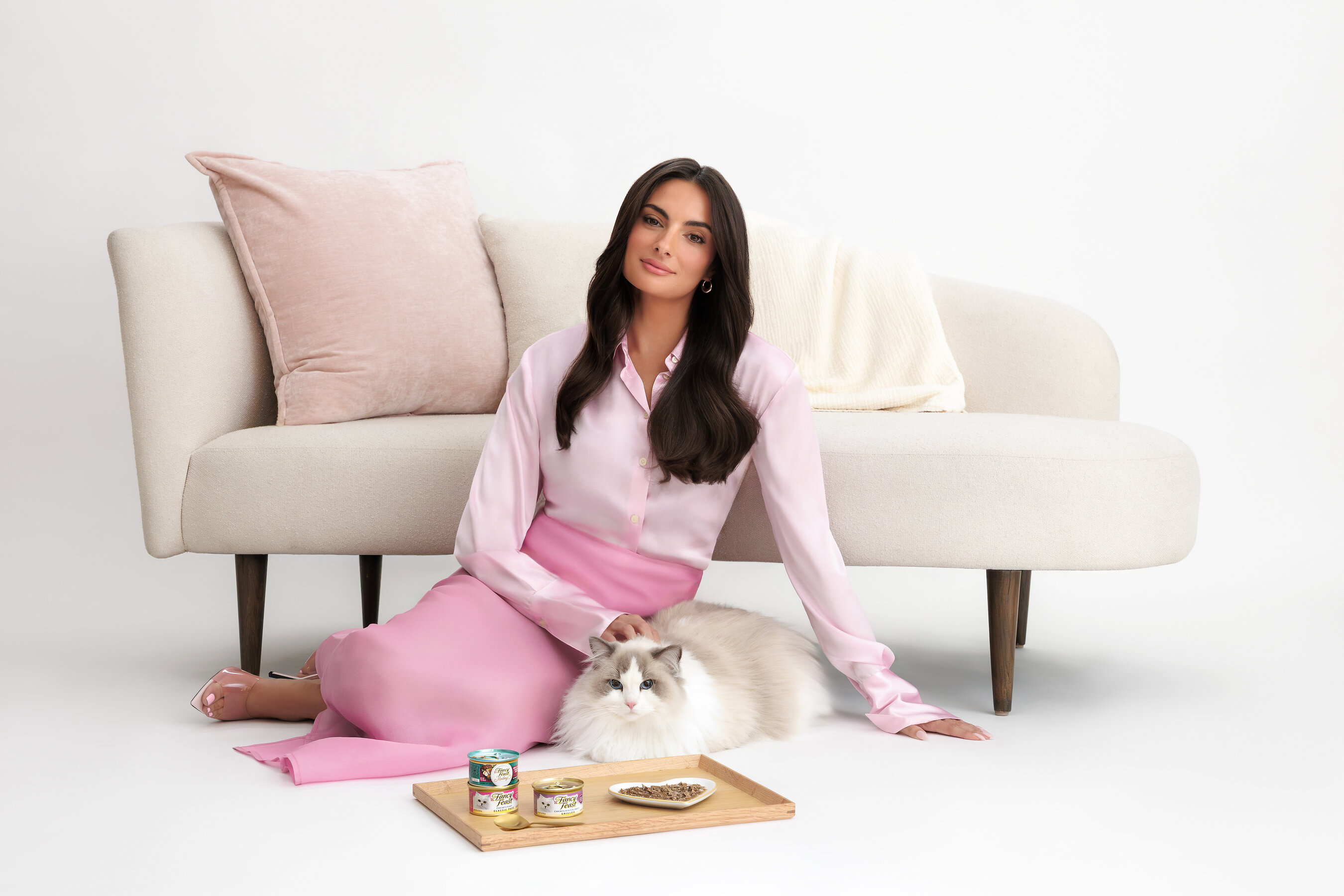 Woman in pink outfit sitting with white cat on cream sofa, pet food products on wooden tray