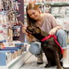 Mulher com chachorro marrom e coleira vermelha no corredor da pet shop testa brinquedo com ele