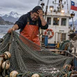 Fisherman on Chilean vessel mending nets with snowy mountains and cat nearby
