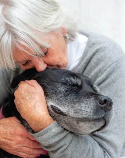 Senior woman with white hair tenderly kissing an older black dog in a warm, affectionate moment