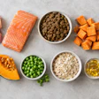 Nutritious whole foods including salmon, chicken, blueberries, sweet potato, carrots, oats, peas, and olive oil arranged on a gray surface