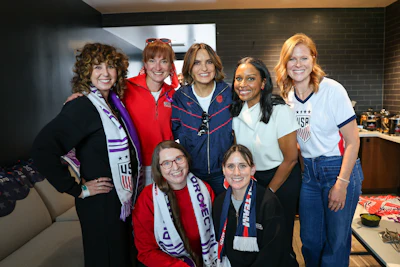 Seven women smiling together at an indoor event, some wearing USA team apparel and medals